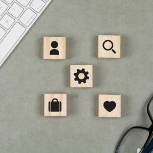 Cinq blocs de bois posés sur un bureau, chacun portant une icône différente : personne, loupe, engrenage, mallette et cœur. Une partie d'un clavier et une paire de lunettes sont également visibles sur les côtés.