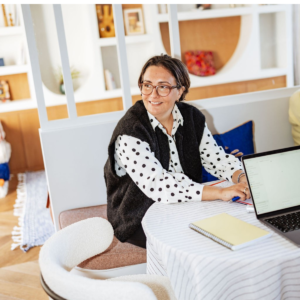 Une personne portant des lunettes, un gilet noir et une chemise à pois est assise à une table avec un ordinateur portable et un bloc-notes, souriant et regardant sur le côté dans un bureau moderne et lumineux.
