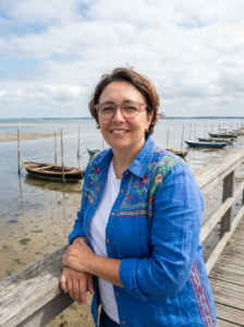 Une femme portant des lunettes et une chemise bleue brodée sourit en s'appuyant sur une balustrade en bois au bord de l'eau, avec de petits bateaux et un ciel partiellement nuageux en arrière-plan.